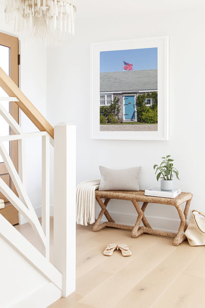 Color fine art photograph of the Wharf Rat Club on Nantucket — weathered grey shingle cottage with a powder blue door, climbing vines, and an American flag flying above the roofline against a blue summer sky