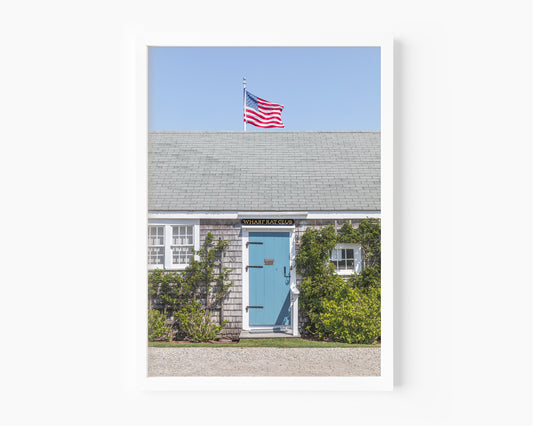 Color fine art photograph of the Wharf Rat Club on Nantucket — weathered grey shingle cottage with a powder blue door, climbing vines, and an American flag flying above the roofline against a blue summer sky