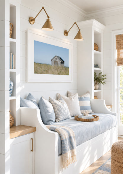 Color fine art photograph of a weathered grey shingle beach cottage alone in golden dune grass on Nantucket Island — ocean visible on the horizon under a wide pale blue sky
