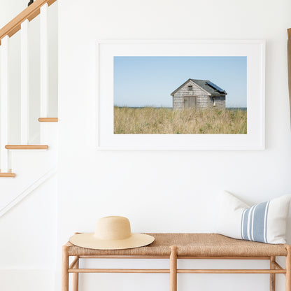 Color fine art photograph of a weathered grey shingle beach cottage alone in golden dune grass on Nantucket Island — ocean visible on the horizon under a wide pale blue sky