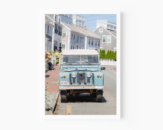 Color fine art photograph of a vintage powder blue Land Rover Series parked on a cobblestone street in Nantucket — Massachusetts vanity plate BLUSKY, grey shingle cottages and hydrangeas in the background