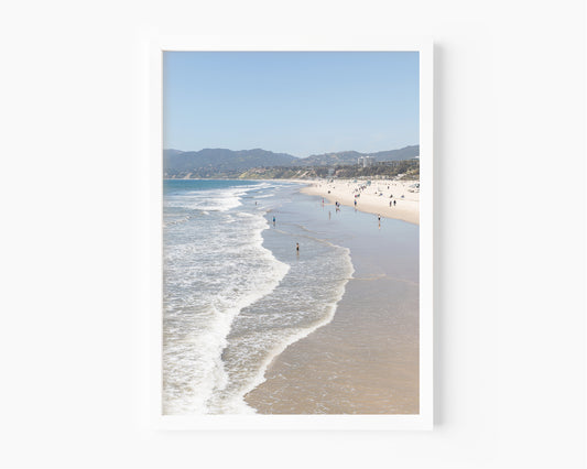 Color fine art photograph of Santa Monica Beach shot from the Santa Monica Pier — rolling Pacific waves, wide sandy shoreline, and the Santa Monica Mountains in the background