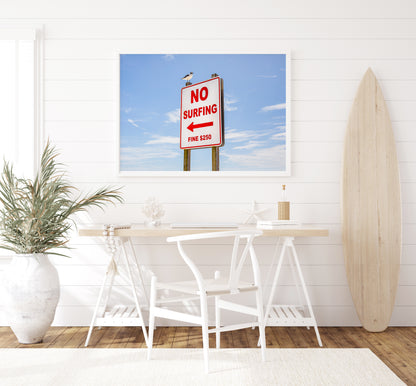 Color fine art photograph of a red and white No Surfing Fine $250 sign at Wrightsville Beach in Wilmington, North Carolina — a seagull perched on top of the sign against a blue sky
