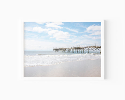 Color fine art photograph of the Ocean Isle Beach fishing pier in North Carolina stretching over the Atlantic — soft waves rolling in along the shoreline under a blue and white sky
