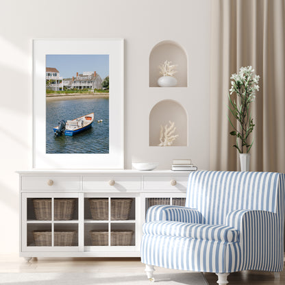 Vertical beach photography print of a small blue boat floating in Nantucket harbor, with Cape Cod-style houses and sandy shoreline in the background — perfect for nautical home decor and coastal wall art.