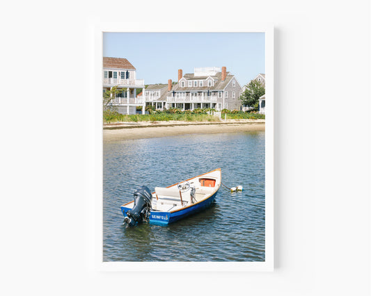 Vertical beach photography print of a small blue boat floating in Nantucket harbor, with Cape Cod-style houses and sandy shoreline in the background — perfect for nautical home decor and coastal wall art.